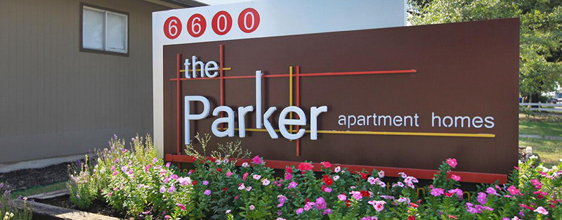 Signboard for 'the Parker apartment homes' with the number 6600, surrounded by flowering plants, in front of a building.