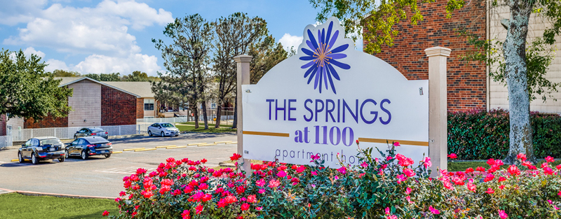 Signage for 'THE SPRINGS at 1100 apartment homes' with a flower logo, surrounded by vibrant pink flowers, in front of the apartment complex with visible parked cars and buildings under a clear blue sky.