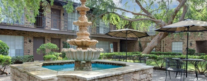 A stone fountain in a courtyard with shaded tables, surrounded by a brick apartment complex with iron railings and mature trees.