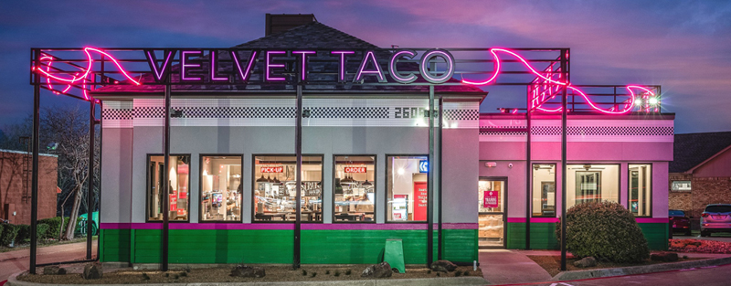 A restaurant at dusk with vibrant neon signage reading 'VELVET TACO', a green and white facade, and visible interior lights.