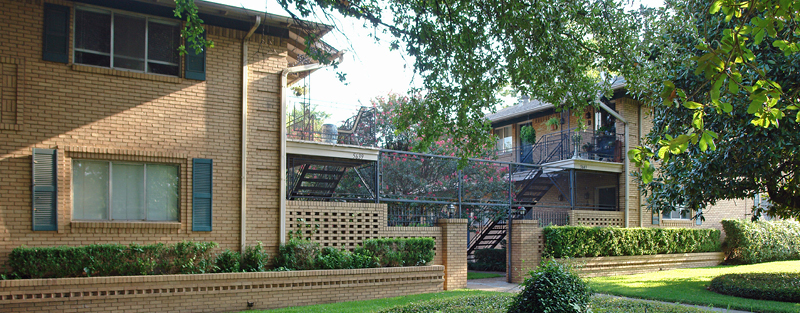 Two-story brick apartment building with external staircases, balconies, and landscaped greenery under a canopy of trees.