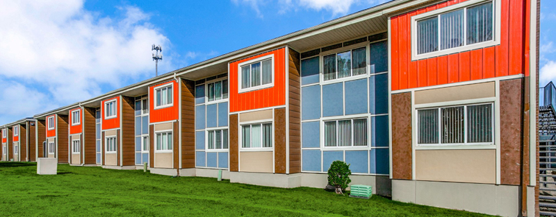 Colorful two-story multifamily housing with red and brown panels, large windows, and an external staircase on a grassy lawn.