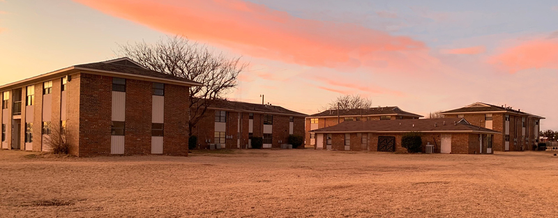 Brick apartment buildings at dusk with a large open lawn and a pinkish sky in the background.