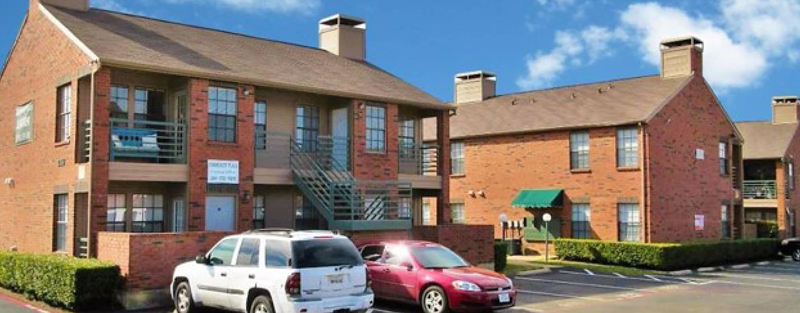 Exterior view of Community Place Apartments with red brick buildings, green stair railings, and parked cars in front.