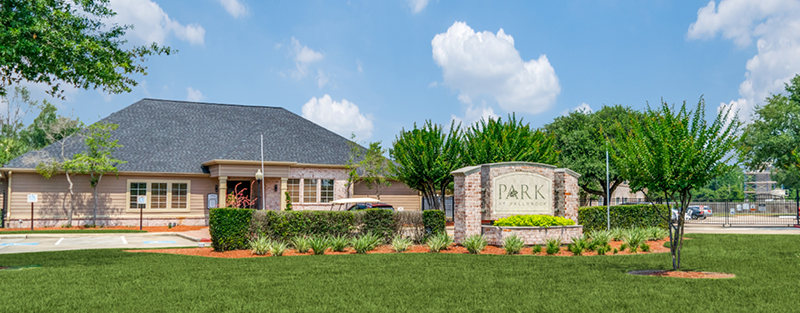 Entrance to Park at Fallbrook Apartments with a brick sign, well-manicured landscaping, and a large single-story building in the background.