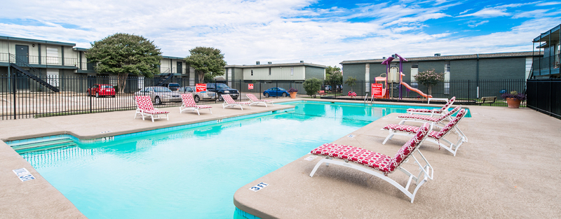 Outdoor swimming pool with red and white lounge chairs, surrounded by a fence, in a residential apartment complex.