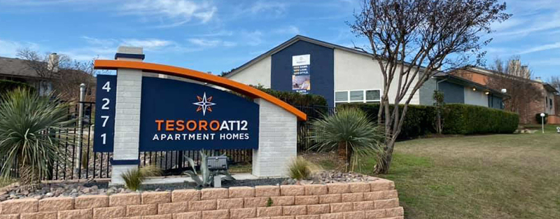 Entrance sign for Tesoro at 12 Apartment Homes with the address 4271, surrounded by landscaping and a view of the apartment buildings in the background.