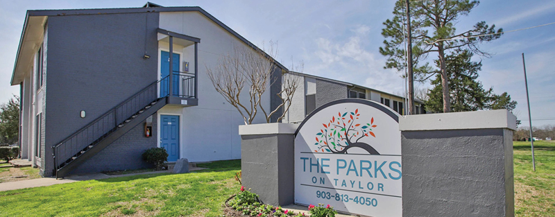 A two-story apartment building with blue doors and a sign in the foreground reading 'The Parks on Taylor,' set in a grassy area with a few trees.