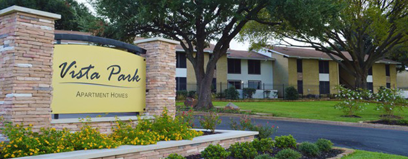 Entrance sign for Vista Park Apartment Homes with landscaped flowers and trees, and a view of the apartment buildings in the background.
