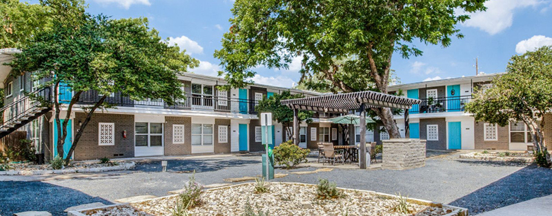 Two-story apartment complex with blue and white accents, featuring a central courtyard with a shaded seating area and mature trees.