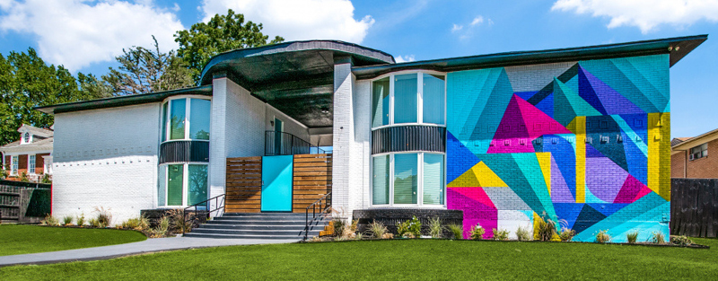 Modern two-story building with large windows, white brick exterior, and a colorful geometric mural on one side.
