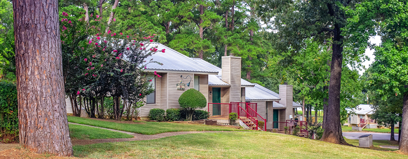 Small residential buildings with metal roofs and front porches, surrounded by lush greenery and tall trees, in a well-maintained landscaped area.