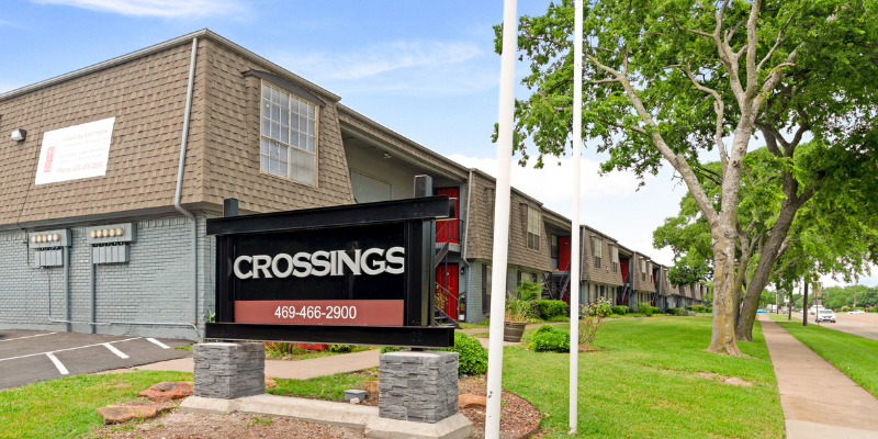 'CROSSINGS' signboard with contact details in front of a brick building on the left, and a sidewalk beside a grassy area with trees lining a street next to apartment buildings on the right.