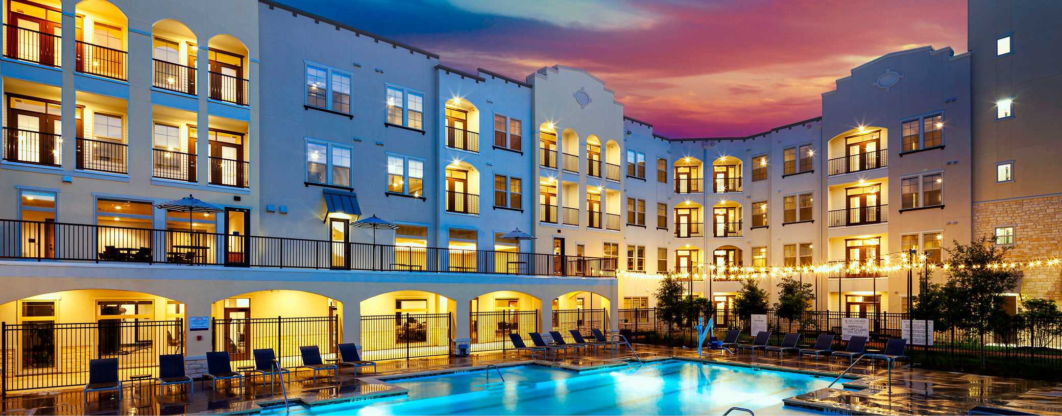 Four-story apartment complex with arched balconies and illuminated windows surrounding a central outdoor swimming pool area under string lights at dusk.