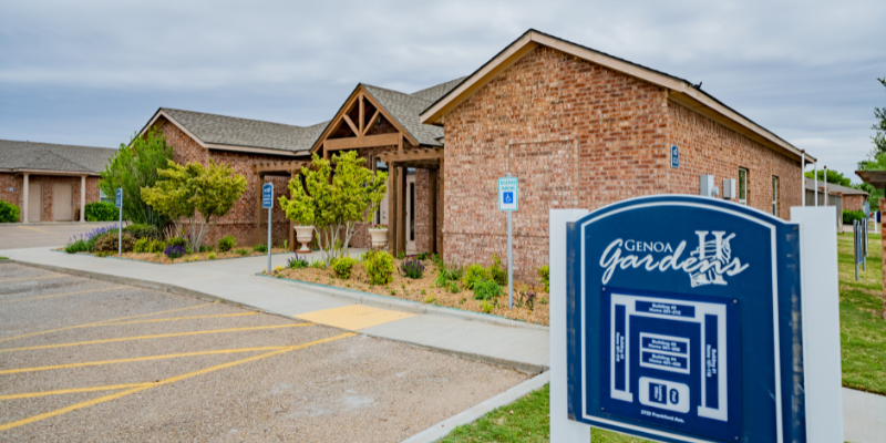 Entrance of 'GENOA GARDENS II' with a prominent sign, brick building in the background, landscaped garden, and handicapped parking, under an overcast sky.