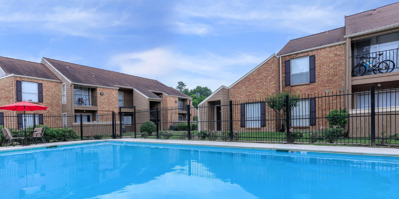 A clear blue swimming pool with lounging chairs and a red umbrella, flanked by two-story brick apartment buildings with balconies.