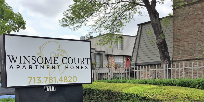 Prominent sign for 'WINSOME COURT APARTMENT HOMES' with contact information in front of a fenced residential area with greenery.