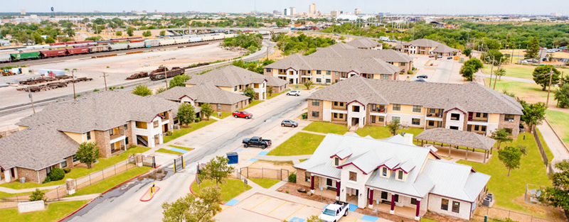 Aerial view of a residential complex featuring multiple two-story buildings with brown roofs, a central office building, and surrounding streets, with a railroad and city skyline in the distance.