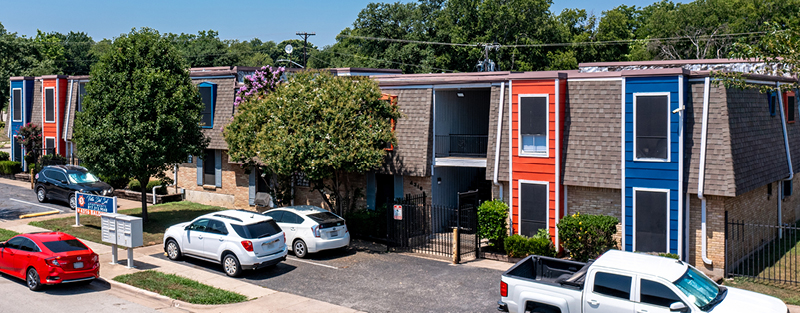 Two-story apartment complex with colorful orange and blue accents, featuring sloped roofs, and a parking area with several vehicles in front.