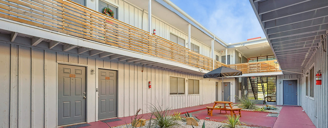 Interior courtyard of a two-story apartment complex with wooden railings, gray doors, red flooring, and a seating area with picnic tables and plants.