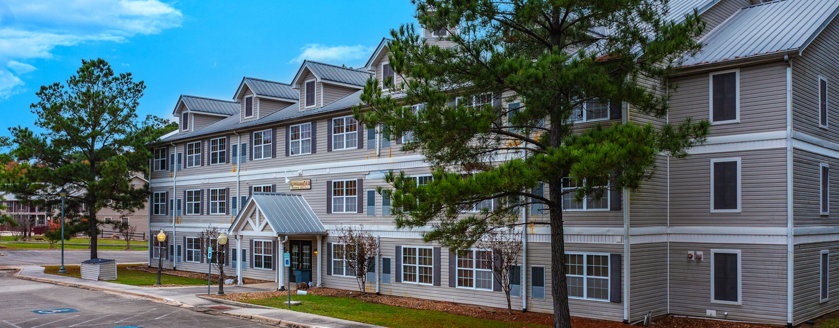 Three-story residential building with gray siding, white trim, dormer windows, and a central entrance, surrounded by trees and a parking area.