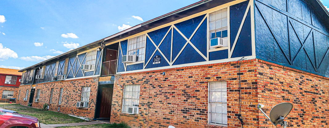 Two-story brick apartment building with blue and white Tudor-style accents, window air conditioning units, and a small grassy area in front.