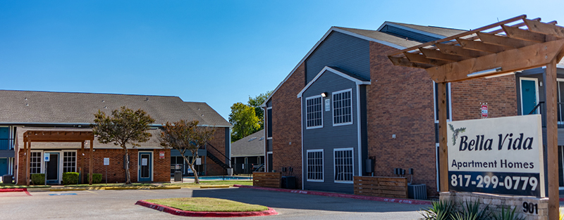 Two-story brick and gray apartment buildings with a sign in front reading 'Bella Vida Apartment Homes' and landscaped areas surrounding the parking lot.