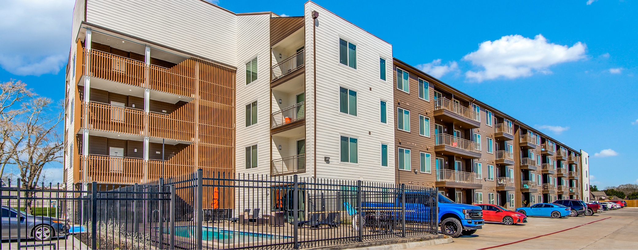 Four-story apartment complex with a mix of white and brown siding, featuring balconies and a fenced pool area, with a parking lot in front and a clear blue sky above.