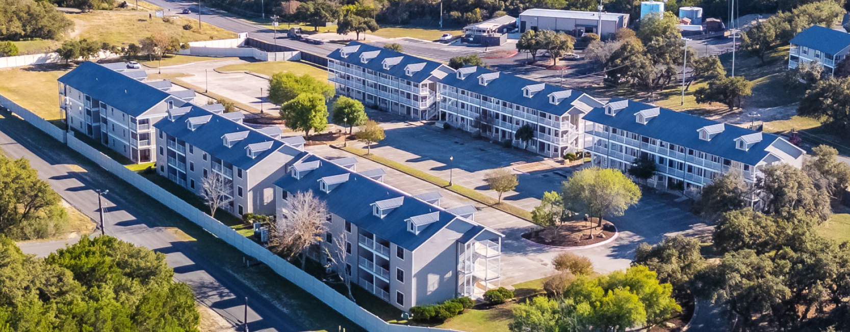 Aerial view of Hill Country Resort, featuring multiple three-story buildings with pitched roofs, surrounded by trees and open parking areas in a Texas hill country setting.