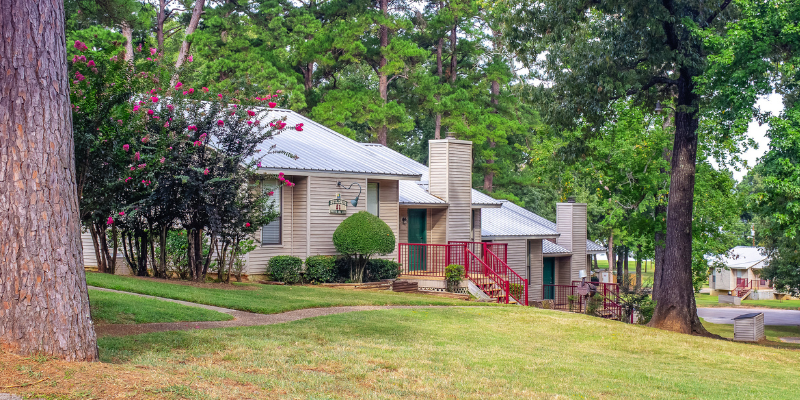 Small residential buildings with metal roofs and front porches, surrounded by lush greenery and tall trees, in a well-maintained landscaped area.