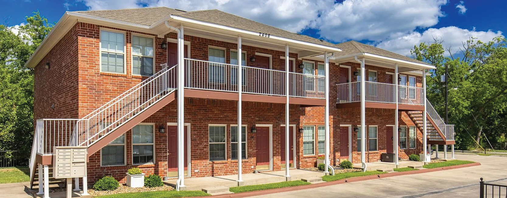 Two-story red brick apartment building with white metal staircases, balconies, and maroon doors, surrounded by greenery and a paved parking area.