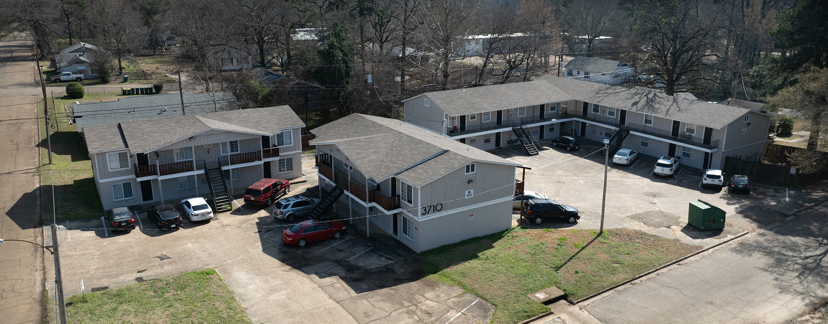 Aerial view of a two-story apartment complex with gray exteriors and pitched roofs, featuring exterior staircases, parking areas, and surrounding trees in a suburban setting.