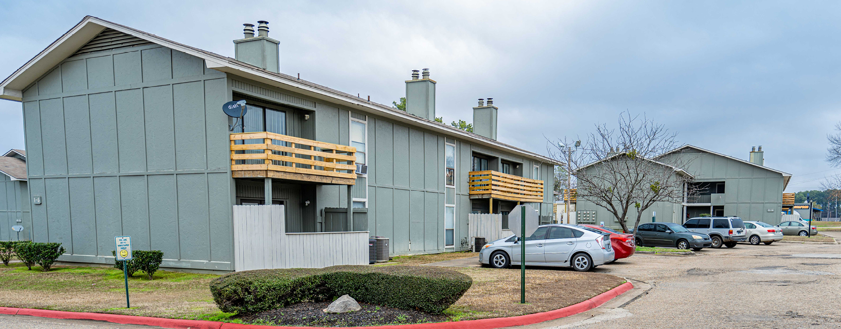 Two-story apartment complex with gray exteriors, wooden balconies, and chimneys, surrounded by a parking lot with several cars and landscaped greenery.