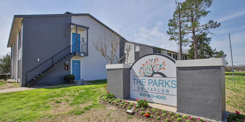 A two-story apartment building with blue doors and a sign in the foreground reading 'The Parks on Taylor,' set in a grassy area with a few trees.