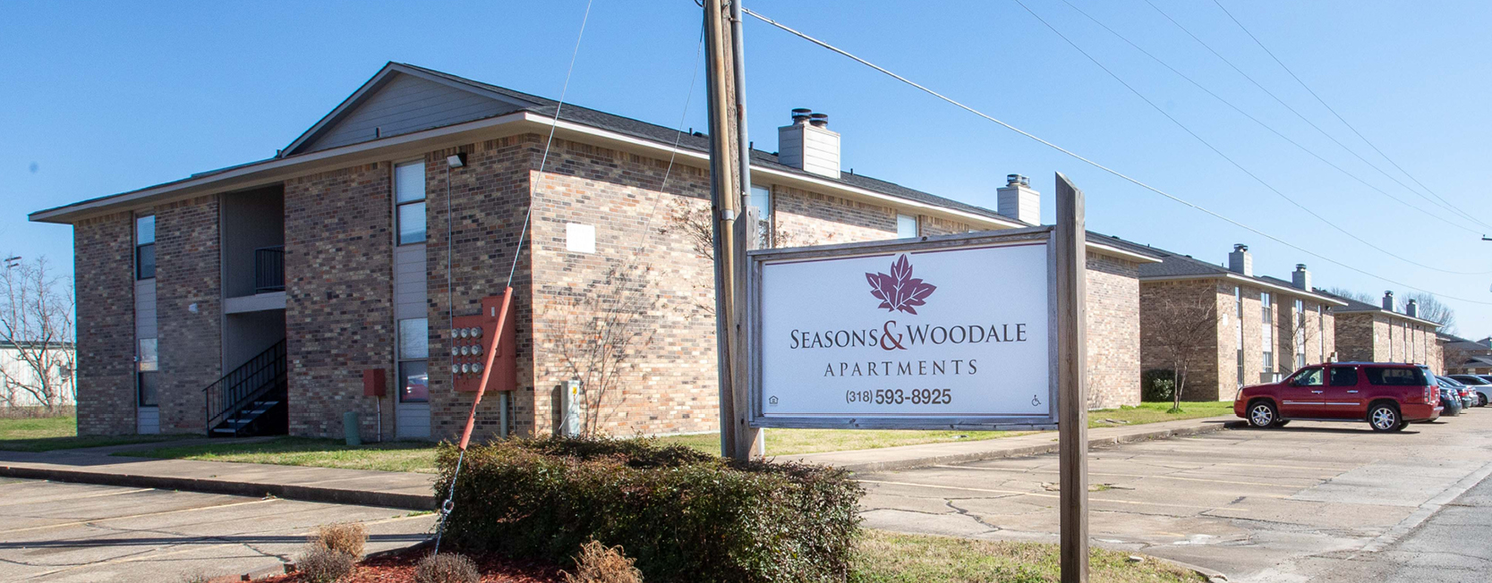 Two-story brick apartment complex with pitched roofs, exterior staircases, and a parking lot, featuring a sign in front reading 'Seasons & Woodale Apartments' with contact information.