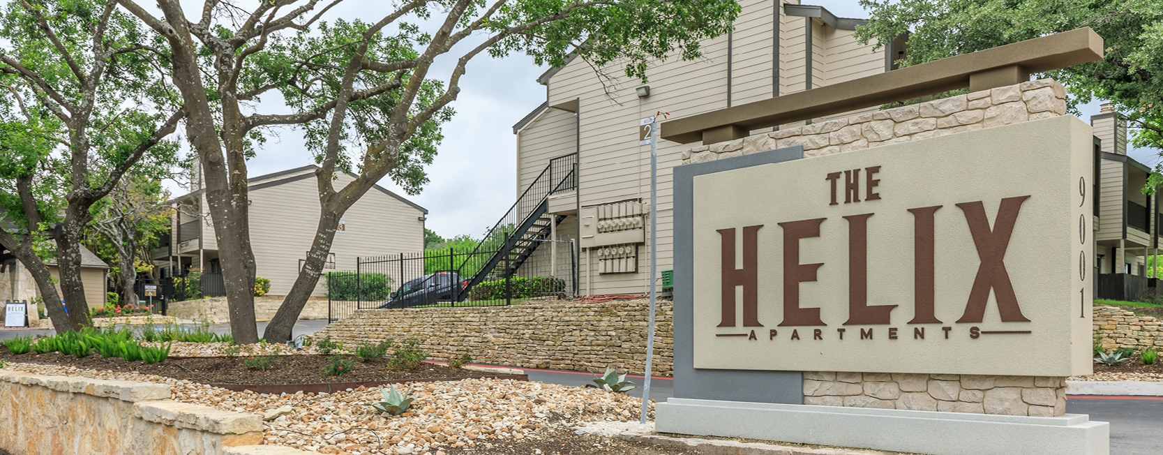 Entrance to The Helix Apartments in San Antonio, TX, featuring a large stone and stucco sign, beige multi-story buildings with exterior staircases, and landscaped areas with trees and rock gardens.