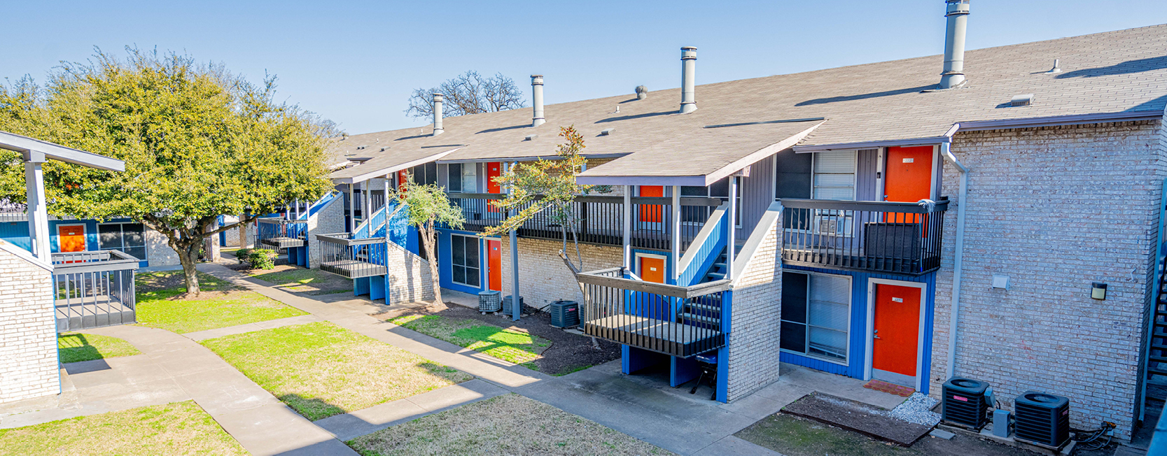 Two-story apartment complex with white brick exteriors, blue railings, and bright orange doors, featuring outdoor staircases, balconies, and a landscaped courtyard with walkways and trees.