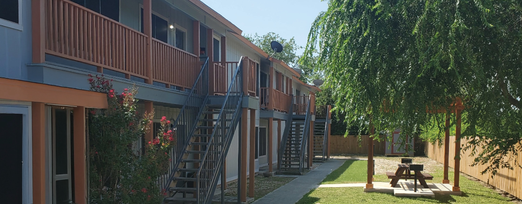 Two-story apartment complex with orange and gray exterior, featuring metal staircases and wooden balconies, alongside a shaded picnic area with BBQ grills and a well-maintained lawn, enclosed by a wooden fence and trees.