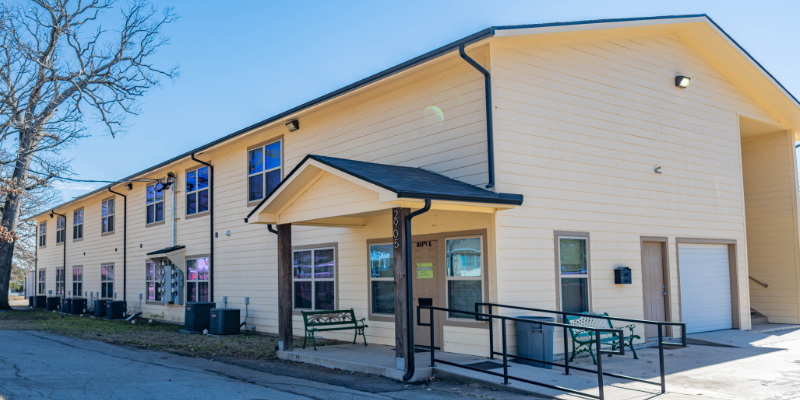 Two-story beige multifamily building with black trim and a covered entrance, adjacent to a bare tree and a clear blue sky.