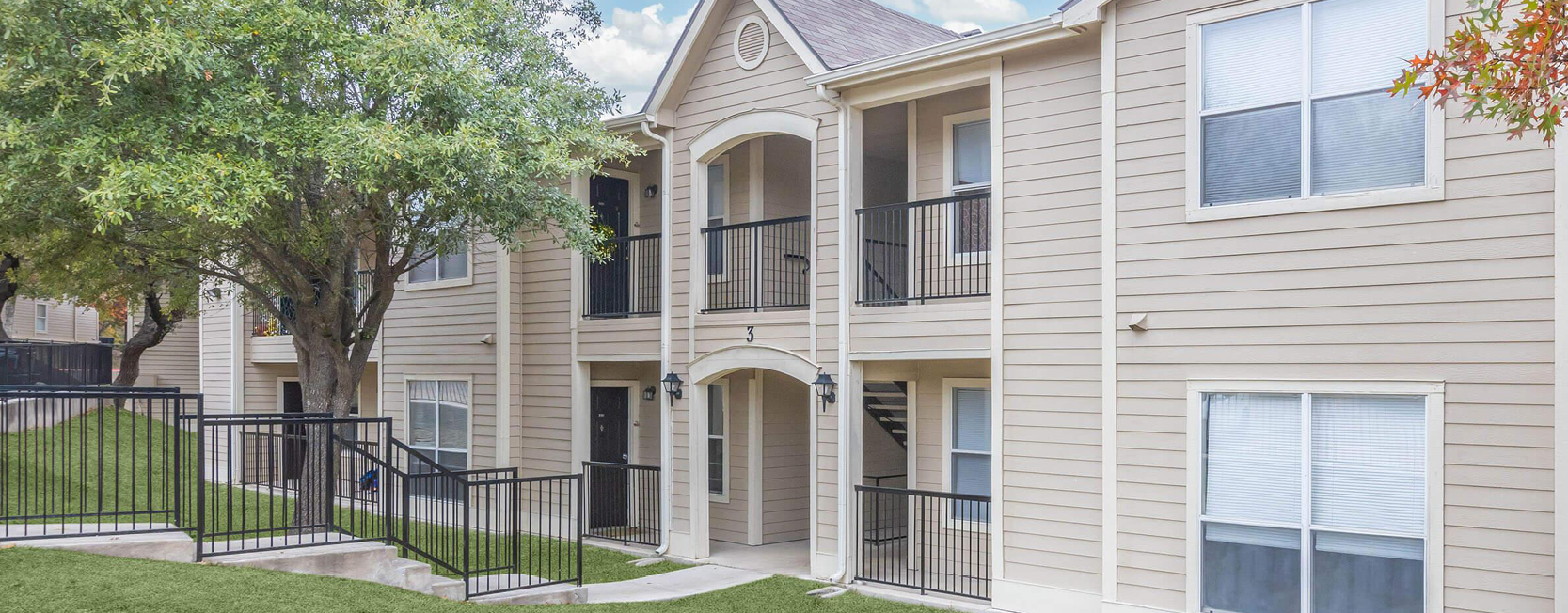 Two-story beige apartment building with white trim and black metal railings, featuring arched entryways and staircases leading to upper units, surrounded by green lawns and mature trees.