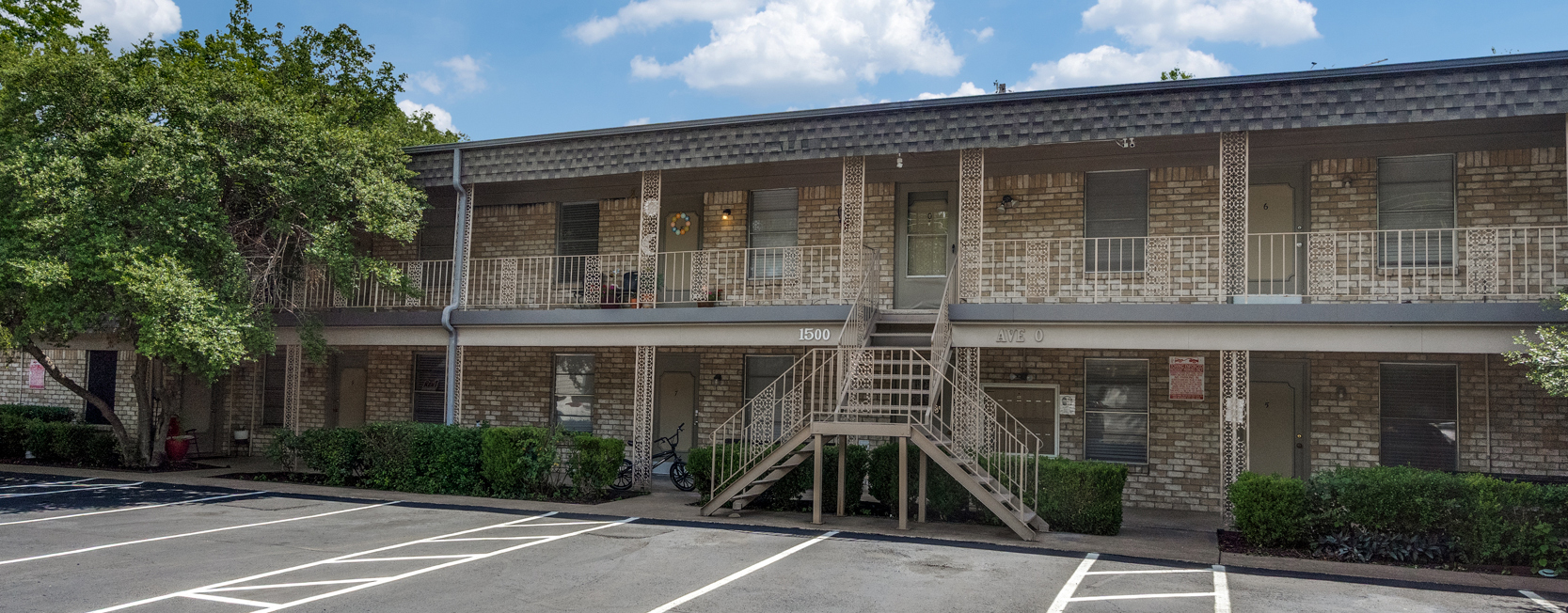 Two-story brick apartment building with balcony railings and central staircase. Address '1500 Ave O' is displayed above the entrance. Empty parking spaces line the front with a few bikes near the units and bushes neatly trimmed along the foundation.