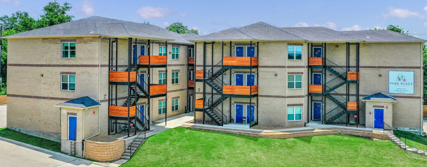 Modern three-story apartment complex with light brick exterior, bold blue doors, and vibrant orange balcony railings. Exterior staircases connect units, and a green lawn stretches across the front courtyard. Sign reads 'Park Place' with contact information.