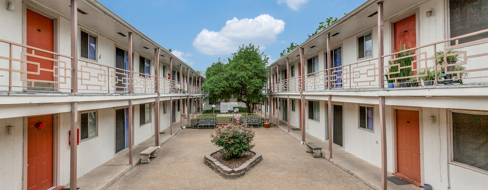 Two-story apartment complex with cream exterior and coral red doors, centered around a landscaped courtyard with rose bushes and benches. Upper-level units feature decorative metal railings and potted plants along the walkway.