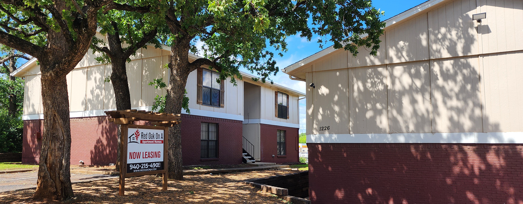 Exterior view of Red Oak On A apartment buildings with two-toned siding (cream and red brick), large mature trees casting shadows, and a wooden 'Now Leasing' sign with contact details out front.