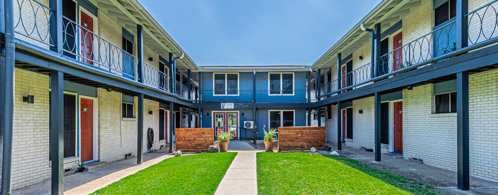 Two-story apartment courtyard with cream brick exterior, red doors, blue railings, and a central lawn leading to a leasing center with a blue facade.