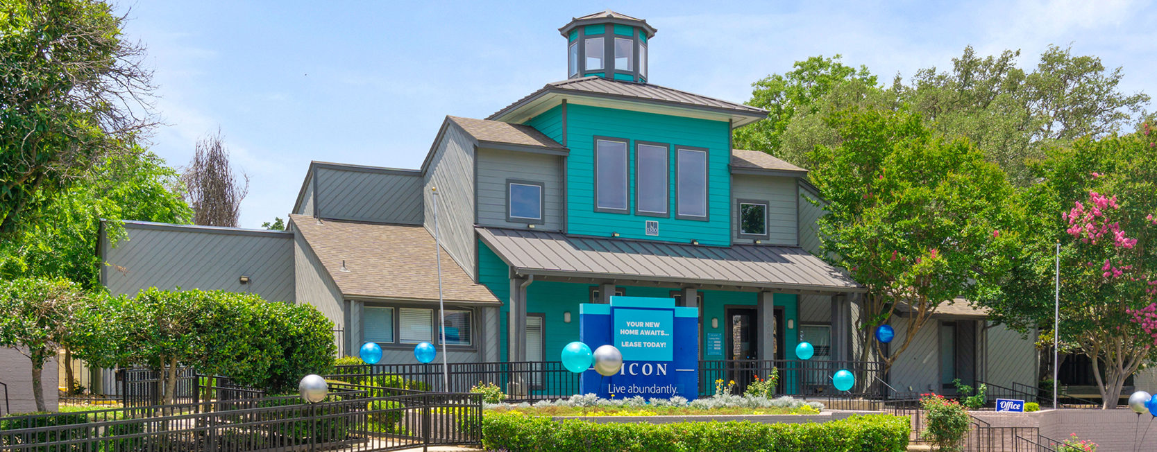 Exterior view of ICON Apartments in San Antonio, TX, featuring a vibrant teal and gray leasing office with a tower-style design, festive blue balloons, and a sign that reads 'Your New Home Awaits... Lease Today!