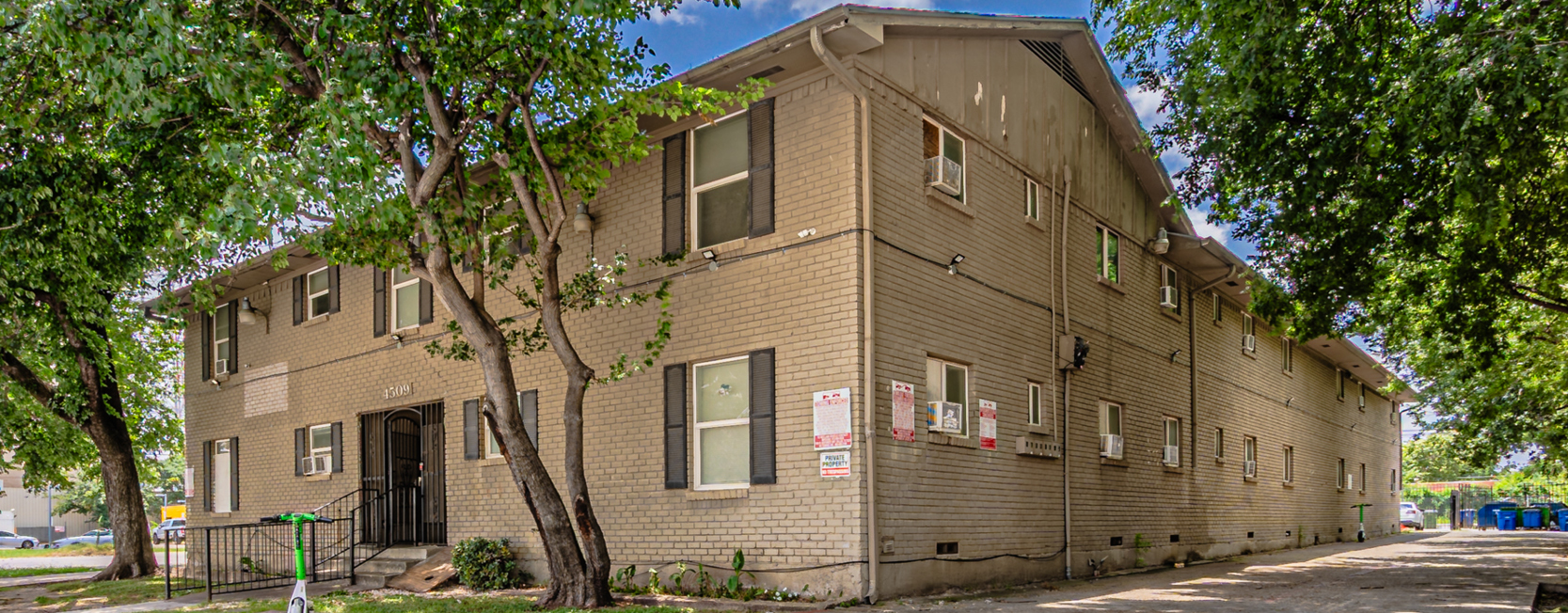 Two-story beige brick apartment complex with dark shutters, exterior window AC units, shaded by mature trees on a residential street.