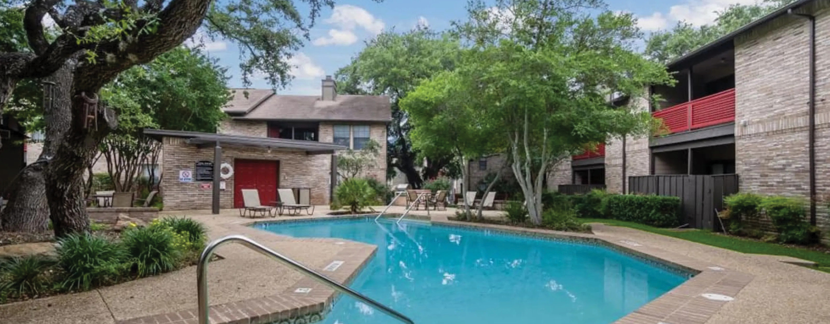 Tranquil pool area surrounded by mature trees, lounge chairs, and two-story brick apartment buildings with red balcony railings.