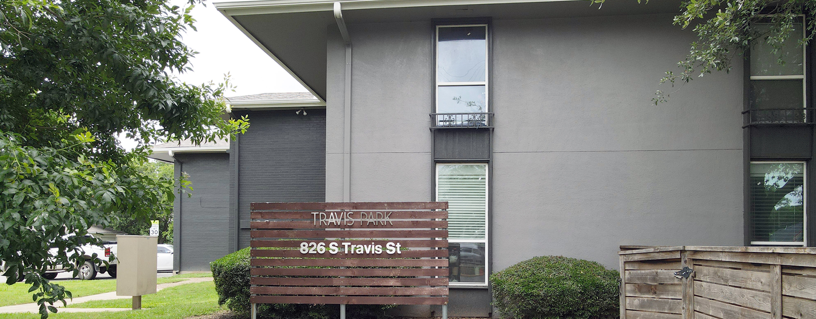 Front view of Travis Park Apartments at 826 S Travis St, featuring a modern gray exterior and a wooden slatted sign with white text, surrounded by greenery and a wooden privacy fence.