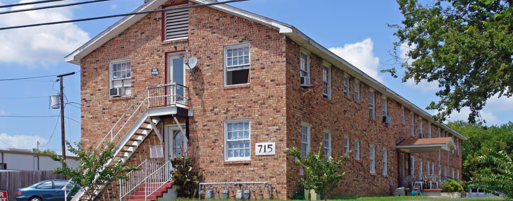 Two-story red brick apartment building with exterior metal stairs, white-trimmed windows, and address number '715' on the front; a modest, older-style multifamily unit in a suburban setting.